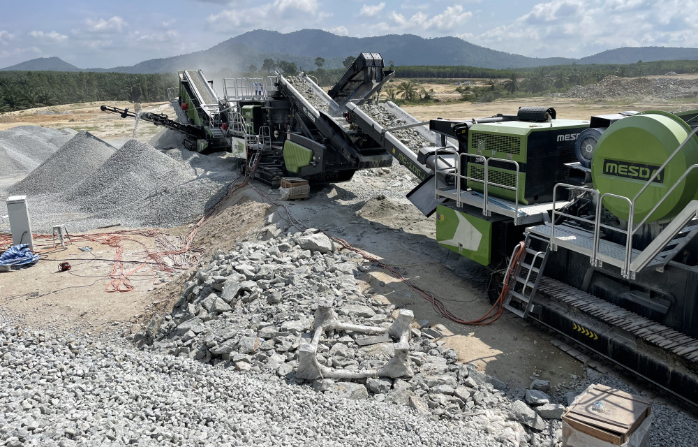 MESDA mobile crusher in operation, discharging crushed aggregate into a large gravel pile at a quarry site with hilly green terrain in the background.